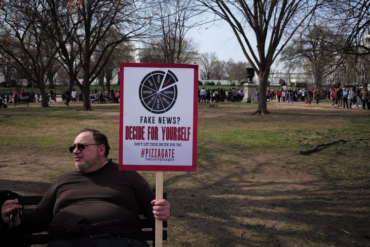 Man sits on a bench holding a sign that says "Fake News? Decide for yourself. #Pizzagate"